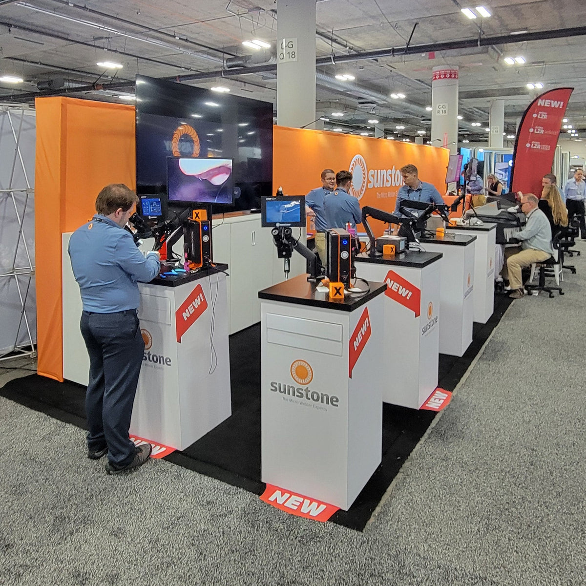 A wide view of a trade show booth area with multiple white and orange display stands, people working at computers, and a large overhead exhibition ceiling with industrial ductwork and lighting.